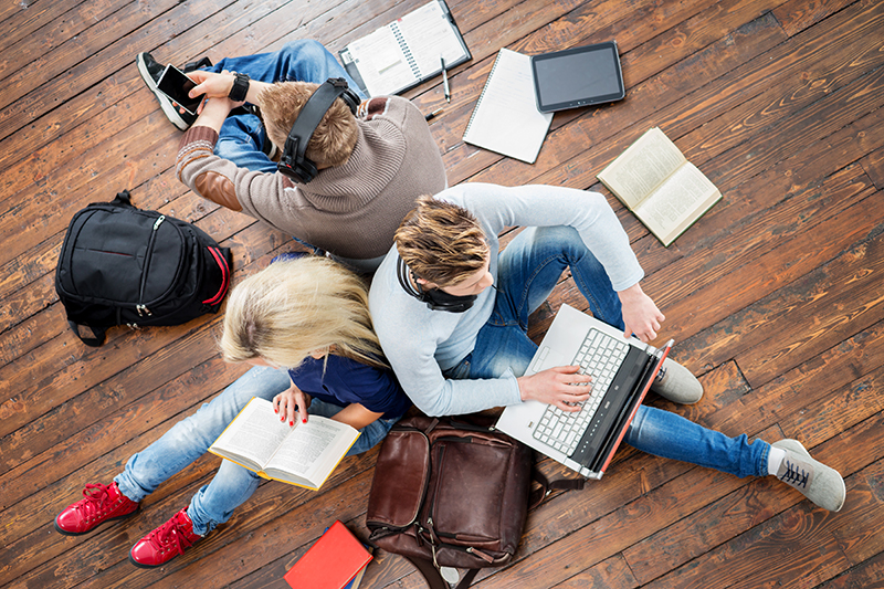 Drei Studierende sitzen auf einem Boden aus Holzdielen und lernen. Ein Studierender trägt Kopfhörer und schaut auf ein Smartphone, ein anderer auf einen Laptop und eine Studierende liest ein Buch. Um sie herum liegen Bücher, Notizbücher und Rucksäcke. Foto: Maksim Shmeljov/colourbox.com