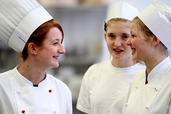 Drei Lehrlinge in weißen Kochjacken und mit Kochmützen während ihrer Ausbildung in der Großküche.