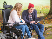 Beispielfoto. Frau im Rollstuhl und ein junger Mann sprechen in einem Park über ein Dokument.
