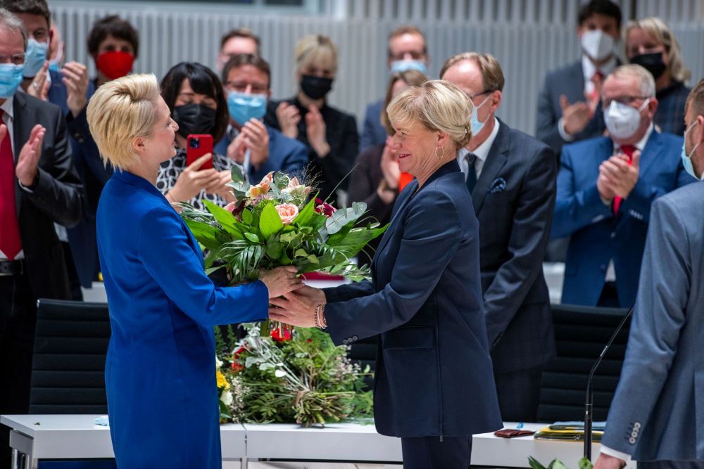 Simone Oldenburg überreicht der frisch gewählten Ministerpräsidentin Manuela Schwesig im Plenarsaal des Landtags einen Blumenstrauß. Im Hintergrund sind applaudierende Abgeordnete zu sehen.