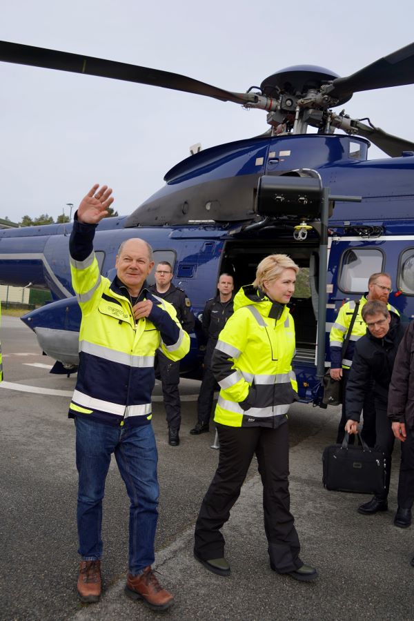 Bundeskanzler Scholz hebt grüßend die Hand. Neben ihm ist Ministerpräsidentin Manuela Schwesig zu sehen. Im Hintergrund steht ein Hubschrauber, vor dem weitere Personen zu erkennen sind.