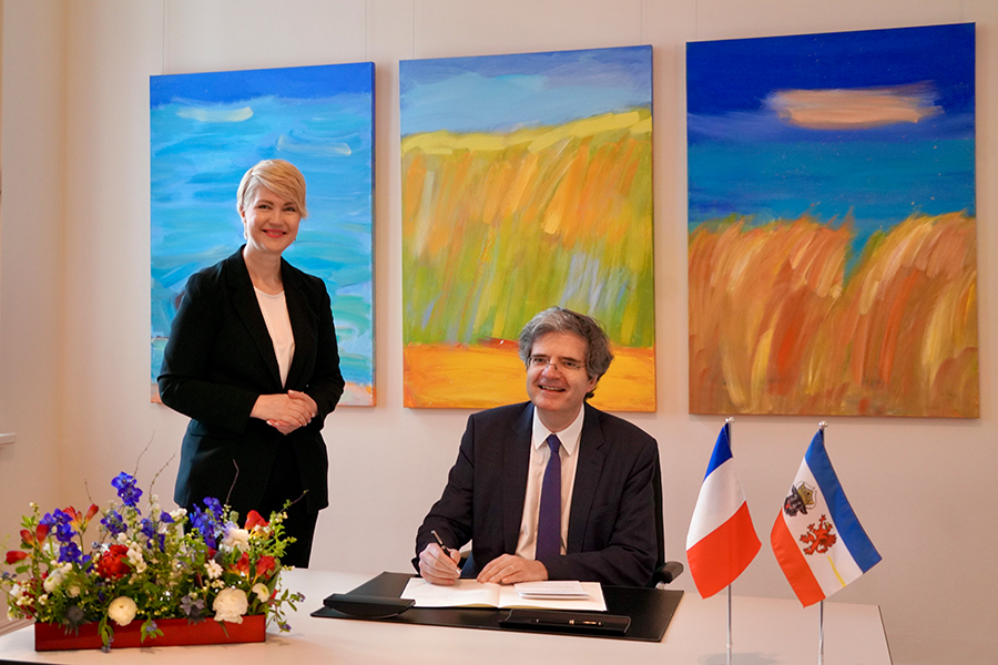 Der französische Botschafter trägt sich ins Gästebuch der Staatskanzlei ein. Die Ministerpräsidentin steht lächelnd hinter dem Tisch. Auf dem Tisch stehen die Flaggen Frankreichs und Mecklenburg-Vorpommerns sowie Blumenschmuck.