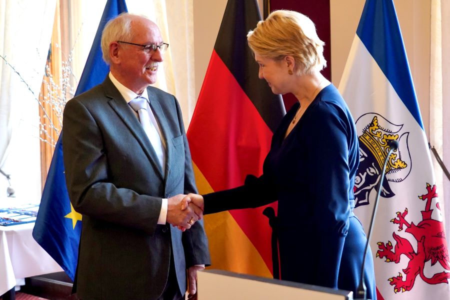 Die Minister­präsidentin gratuliert Heiko Brunner zum Bundes­verdienst­orden. Im Hintergrund sind die EU- und die Deutschland-Flagge sowie die Landesflagge von Mecklenburg-Vorpommern zu sehen. 