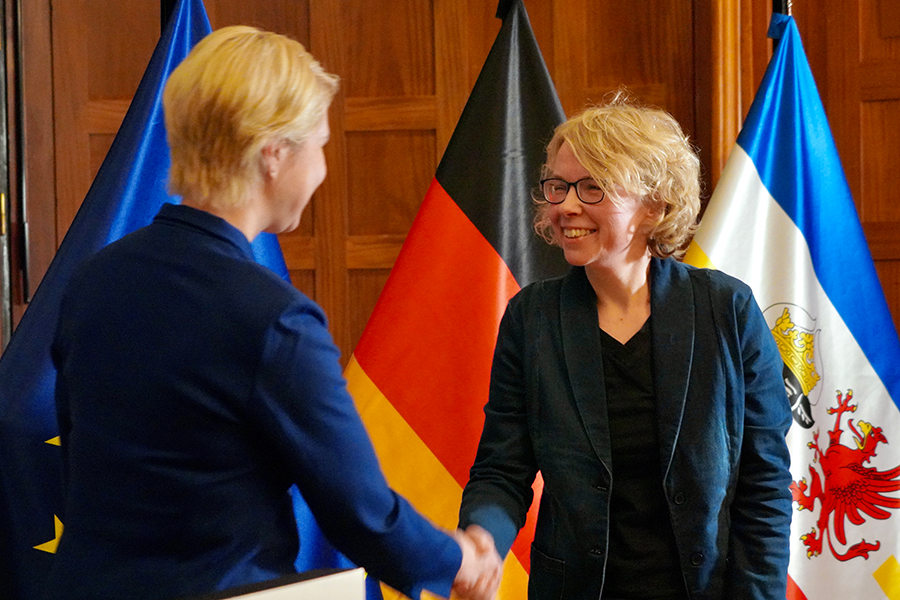 Die Ministerpräsidentin gratuliert Kati Mattutat zu ihrer Auszeichnung. Im Hintergrund sind die EU-, die Deutschland- und die Mecklenburg-Vorpommern-Flagge zu sehen.