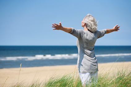 Auf dem Bild steht eine Seniorin mit ausgestreckten Armen und den Kopf in den Wind gestreckt auf einer Düne, hinter ihr ist ein bisschen Gras, vor ihr sieht man Strand und das Meer mit ruhigen Wellen.