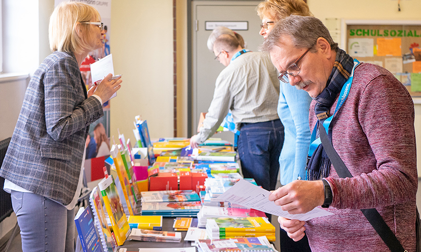 Messetisch mit Schulbüchern, an dem sich eine Vertreterin der Verlage und ein Fortbildungsteilnehmer unterhalten