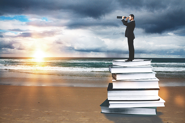 Ein Mann steht auf einem Bücherstapel am Strand und schaut mit einem Fernrohr auf das Meer hinaus. Foto: peshkova/Fotolia.com