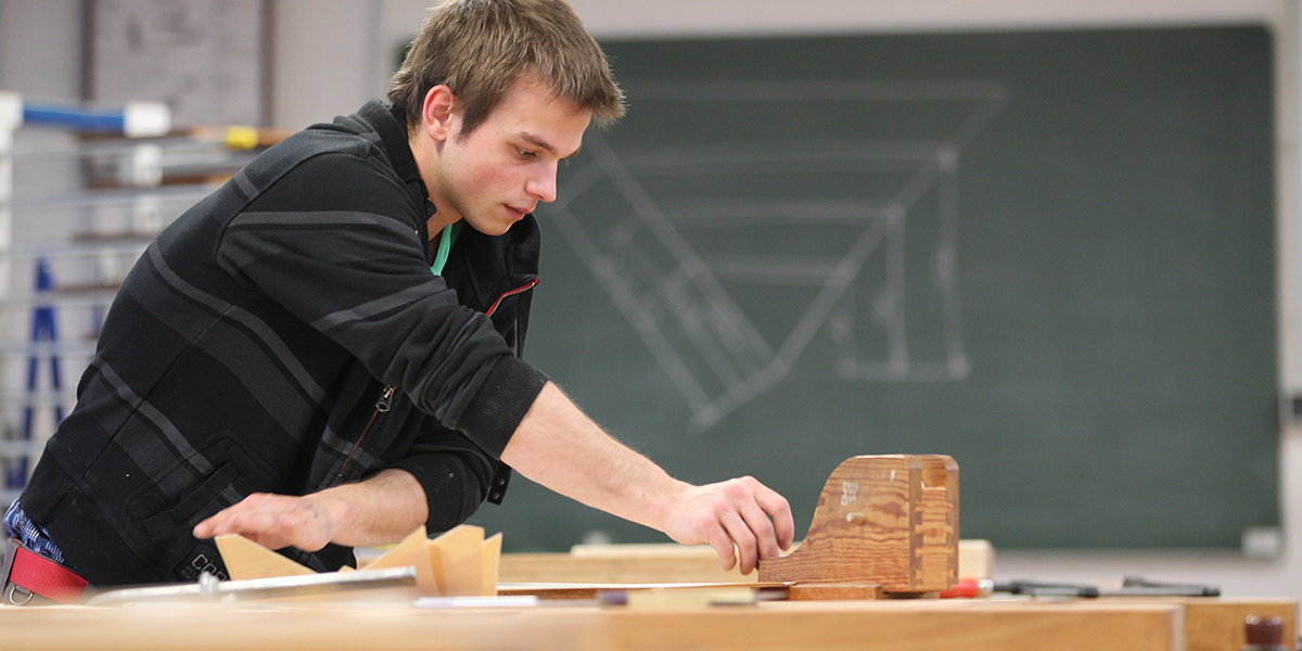 Ein Tischler-Lehrling beugt sich über eine Werkbank, auf der verschiedene Holzteile liegen. Foto: Danny Gohlke