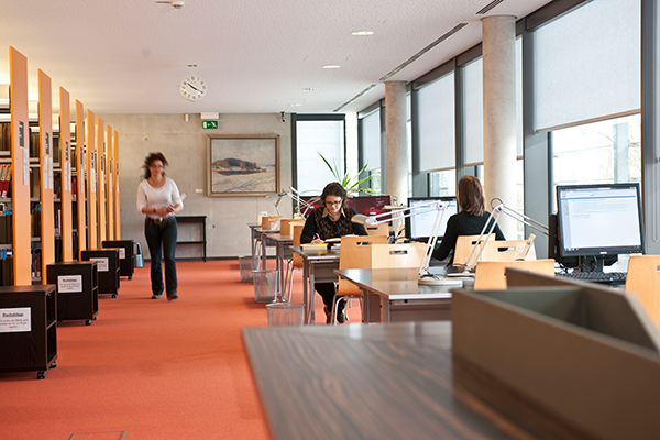 Zwei Frauen sitzen an den Computerarbeitsplätzen in der Landesbibliothek, eine Frau läuft mit einem Buch in den Händen den Gang entlang, links stehen Bücherregale.