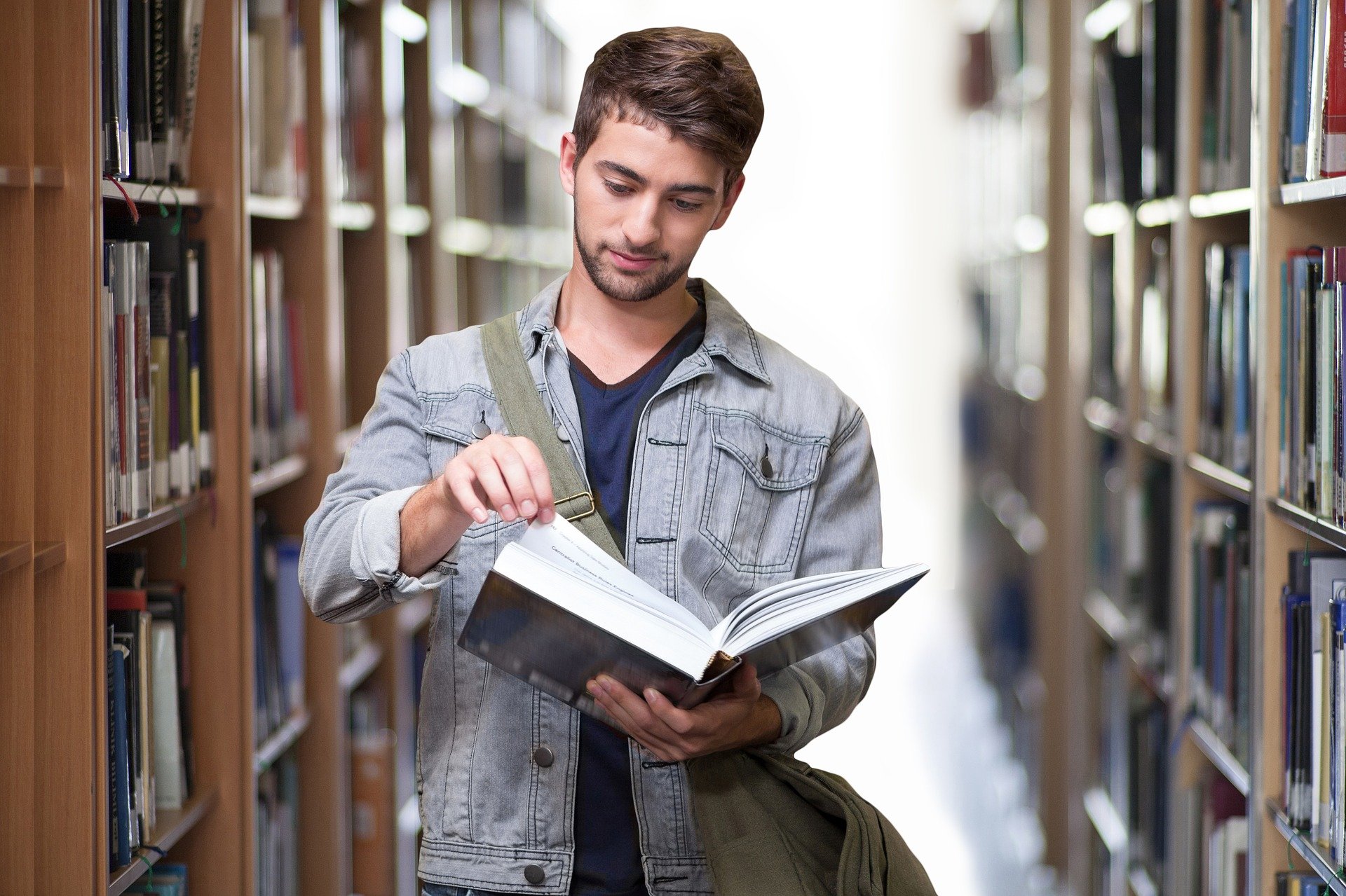 Ein Studierender steht in der Bibliothek zwischen Bücherregalen und blättert in einem Buch. (Interner Link: BAföG)