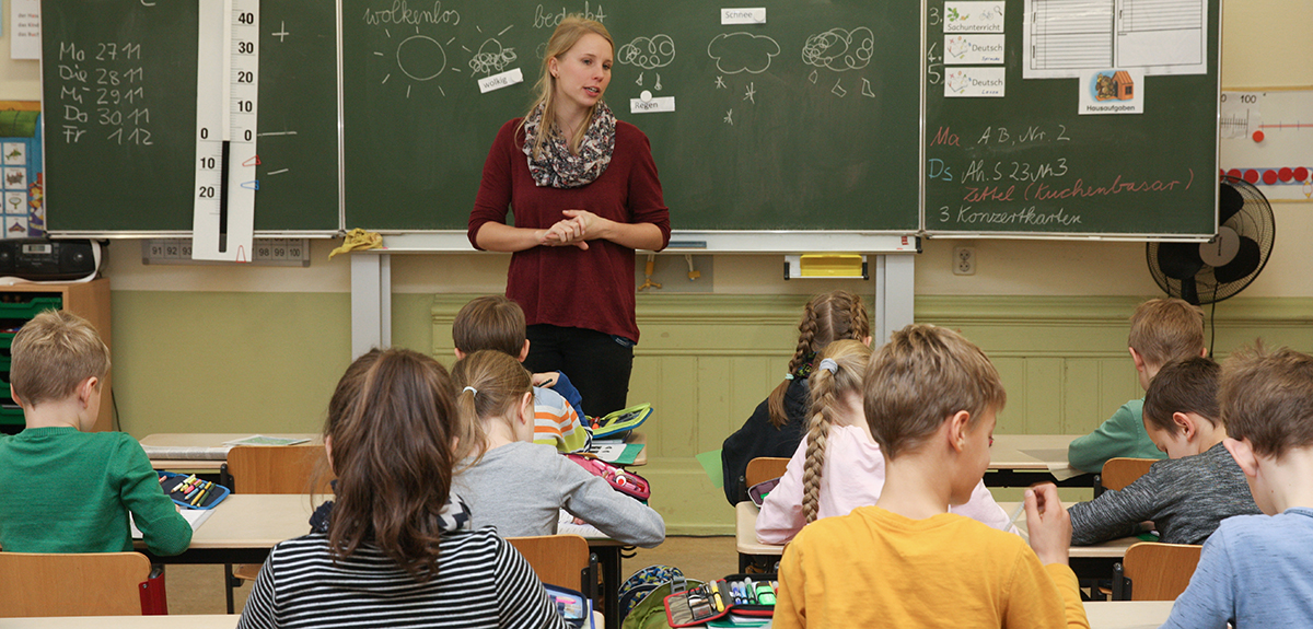 Eine Lehrerin steht an der Tafel und spricht zu ihren Schülerinnen und Schüler, die vor ihr an Tischreihen sitzen. Foto: Silke Winkler