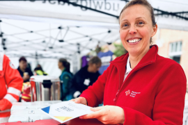 Eine Frau mit roter DRK-Jacke hält lächelnd ein Ehrenamtsdiplom in den Händen und streckt es Richtung Kamera. Im Hintergrund sind verschwommen weitere Menschen.