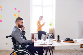 Mann im Rollstuhl von Seite zu sehen sitzt an einem großen Schreibtisch und arbeitet am Laptop