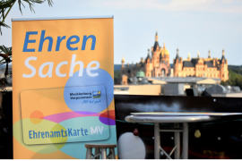 Links ist ein orangenes Banner mit der Aufschrift Ehrensache, darunter ist ein Bild der Ehrenamtskarte MV. Rechts im Bild ist verschwommen das Schweriner Schloss, darüber graublauer Himmel