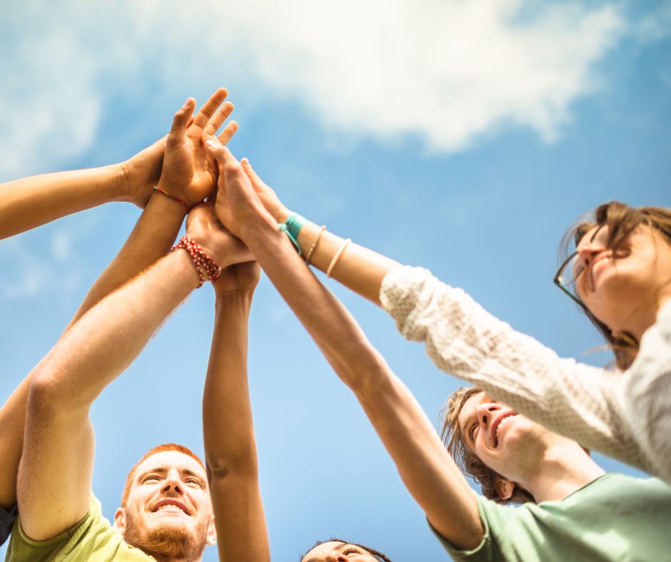 Junge Menschen strecken ihre Arme nach oben, sodass die Hände alle an einem Punkt aufeinander treffen, dahinter ist blauer Himmel mit weißen Wolken