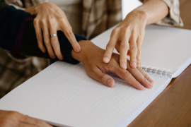 Zwei Hände führen die Hände einer anderen Person über ein Papier mit Braille-Schrift Zwei Hände führen die Hände einer anderen Person über ein Papier mit Braille-Schrift