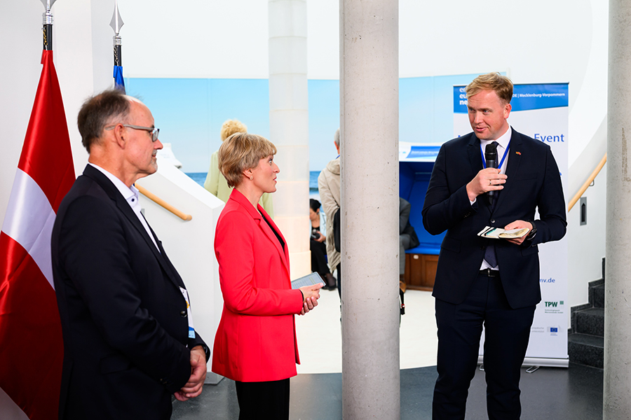 Wirtschaftsminister Viktors Valainis hält ein Mikrophon in der Hand. Zu sehen sind weiter Moderatorin Marianna Evenstein und Wirtschaftsminister Dr. Wolfgang Blank.