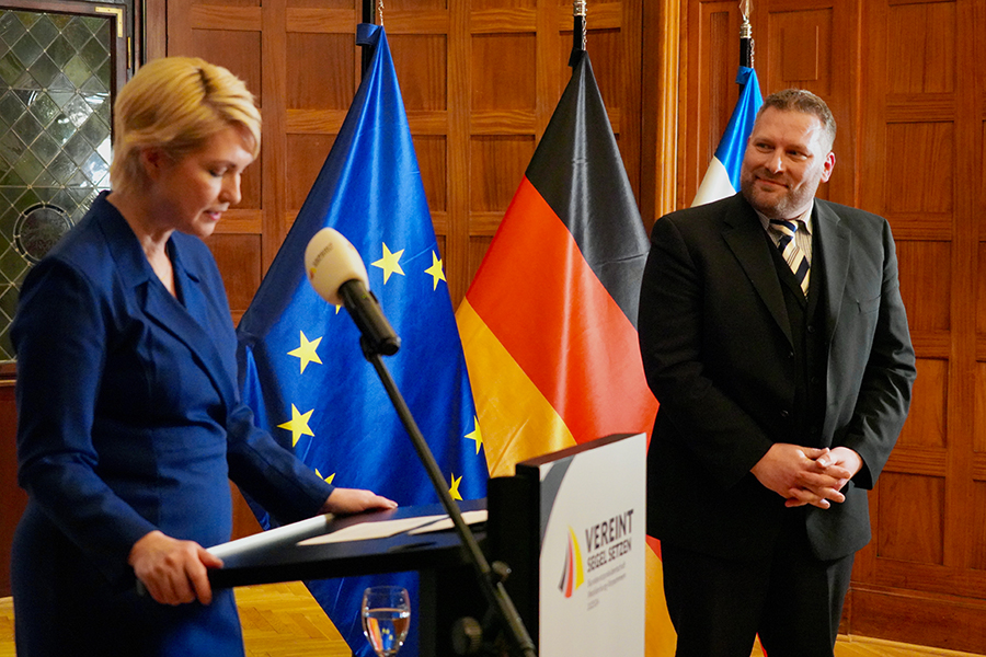 Dr. Daniel Trepsdorf neben dem Redepult, an dem die Ministerpräsidentin seine Verdienste um die Demokratie erläutert. Im Hintergrund sind die EU-, die Deutschland- und die Mecklenburg-Vorpommern-Flagge zu erkennen.