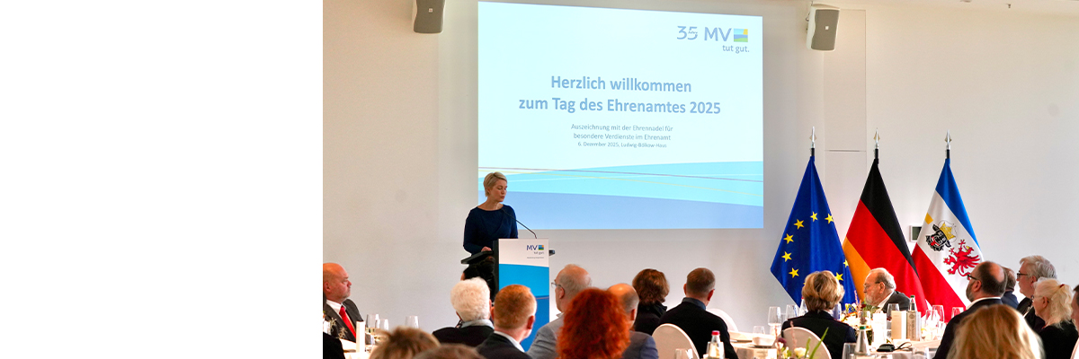 Die Ministerpräsidentin bei ihrer Festrede zum Tag des Ehrenamtes. Im Hintergrund ist ein Bildschirm zu sehen, auf dem  "Herzlich willkommen zum Tag des Ehrenamtes 2025" steht. Im Vordergrund sitzen Gäste an runden Tischen. Rechts sind die EU-Flagge, die Deutschland- sowie die Mecklenburg-Vorpommern-Flagge zu sehen.