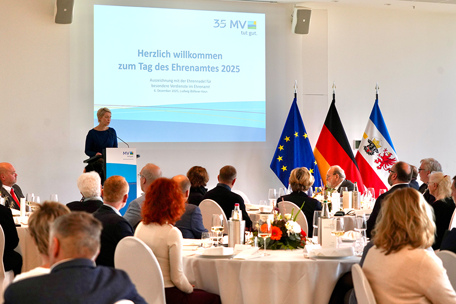 Die Ministerpräsidentin bei ihrer Festrede zum Tag des Ehrenamtes. Im Hintergrund ist ein Bildschirm zu sehen, auf dem  "Herzlich willkommen zum Tag des Ehrenamtes 2025" steht. Im Vordergrund sitzen Gäste an runden Tischen. Rechts sind die EU-Flagge, die Deutschland- sowie die Mecklenburg-Vorpommern-Flagge zu sehen.