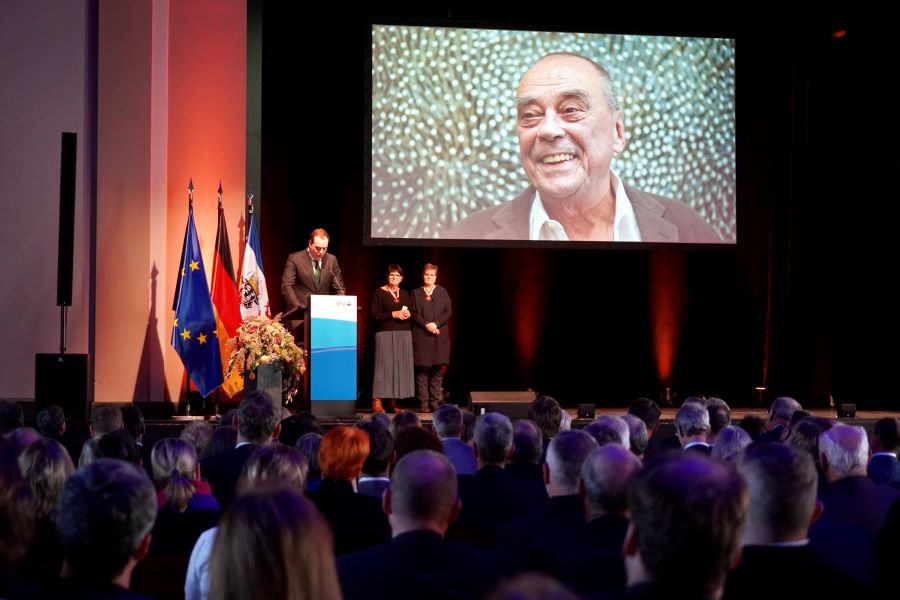 Günther Uecker, auf dem großen Bild im Hintergrund der Bühne, erhielt den Verdienstorden des Landes. Sein Sohn Jacob Uecker steht am Rednerpult und richtet dem Publikum dessen Gruß aus.