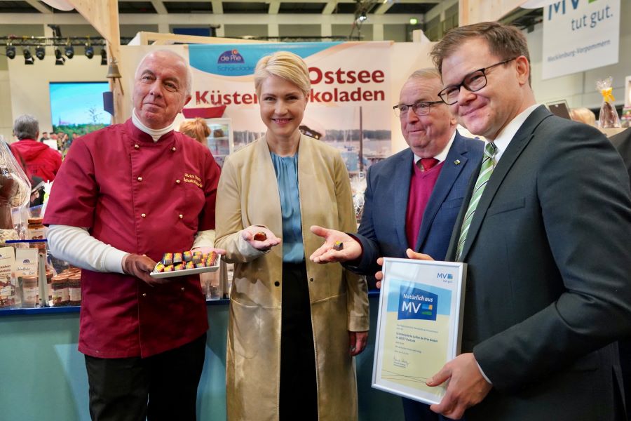 Ministerpräsidentin Manuela Schwesig und Minister Dr. Till Backhaus mit Staatsminister Carsten Schneider, eine Urkunde vorzeigend, und Ulrich Deprie, der ein Tablett mit Pralinen in der Hand hält.