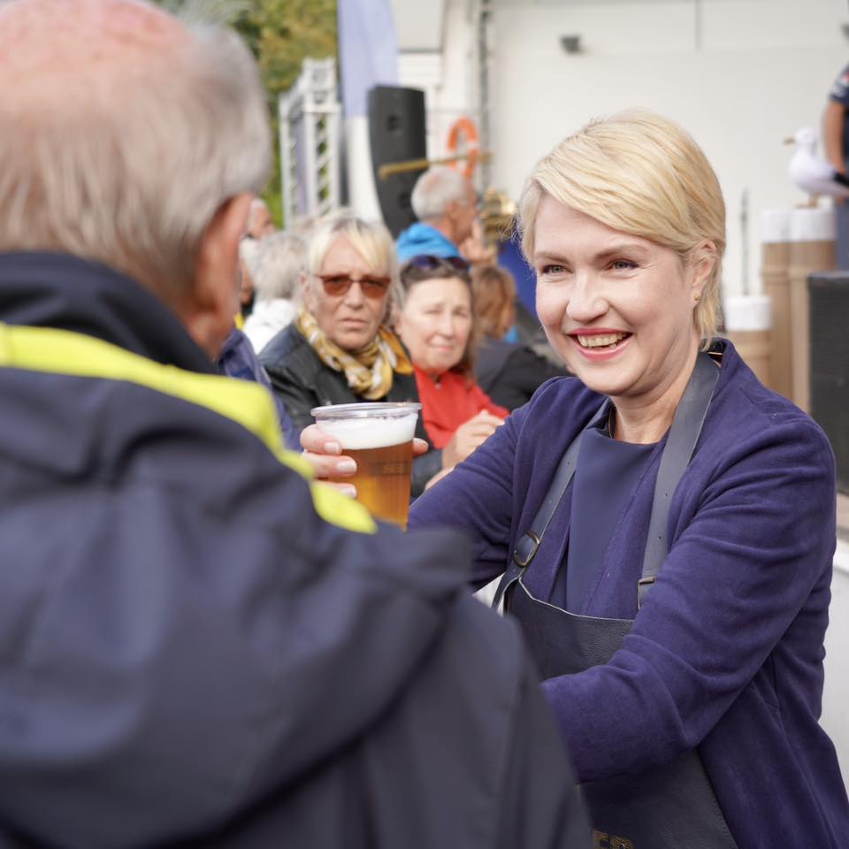 Die Ministerpräsidentin überreicht einem Bürger lächelnd ein Glas Bier. Sie hat eine Schürze um.