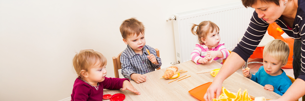 Kinder beim Essen in einer Kita. Eine Erzieherin serviert Obst.