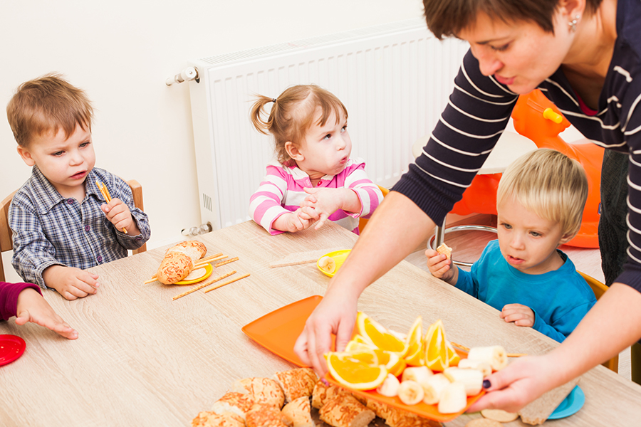Kinder beim Essen in einer Kita. Eine Erzieherin serviert Obst. © Ingram Image, Adpic Kinder beim Essen in einer Kita. Eine Erzieherin serviert Obst.