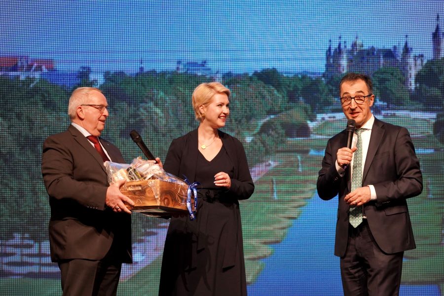 Ministerpräsidentin Manuela Schwesig und Minister Dr. Backhaus überreichen Bundes­landwirt­schafts­minister Cem Özdemir eine Kiste mit Landes­produkten. Im Hintergrund ist ein Foto des Schweriner Schlossgartens mit Blick auf das Schloss zu sehen.