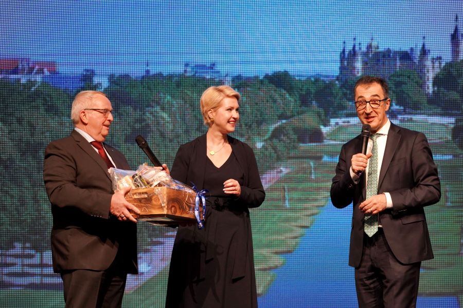 Ministerpräsidentin Manuela Schwesig und Minister Dr. Backhaus überreichen Bundeslandwirtschaftsminister Cem Özdemir eine Kiste mit Landesprodukten. Im Hintergrund ist ein Foto des Schweriner Schlossgartens mit Blick auf das Schloss zu sehen. © StK M-V Ministerpräsidentin Manuela Schwesig und Minister Dr. Backhaus überreichen Bundeslandwirtschaftsminister Cem Özdemir eine Kiste mit Landesprodukten. Im Hintergrund ist ein Foto des Schweriner Schlossgartens mit Blick auf das Schloss zu sehen.