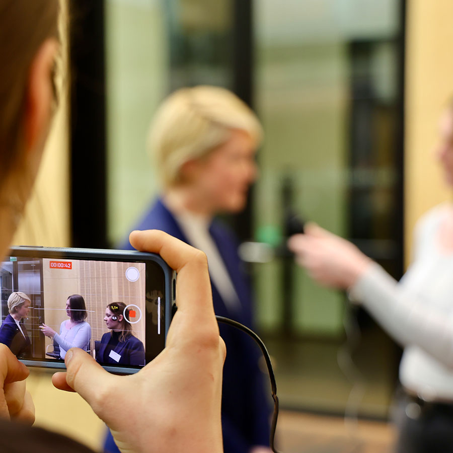 Die Ministerpräsidentin ist auf einem Handybildschirm während eines Video-Interviews zu sehen, Dahinter ist die Originalszene zur erkennen.