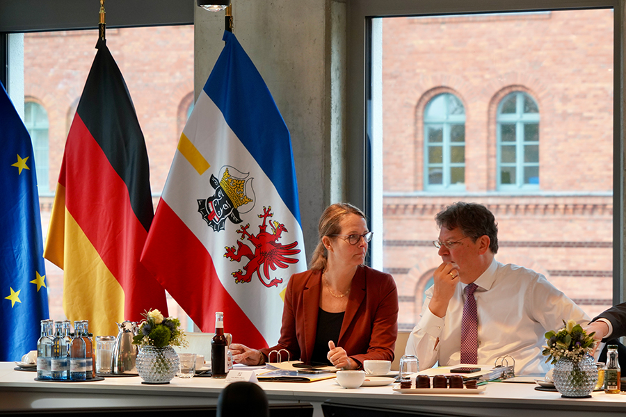 Ministerin Bettina Martin im Gespräch mit Minister Reinhard Meyer am Kabinettstisch. Im Hintergrund sind vor den Fenstern die Flaggen von Mecklenburg-Vorpommern, der Bundesrepublik Deutschland sowie der Europäischen Union zu sehen.