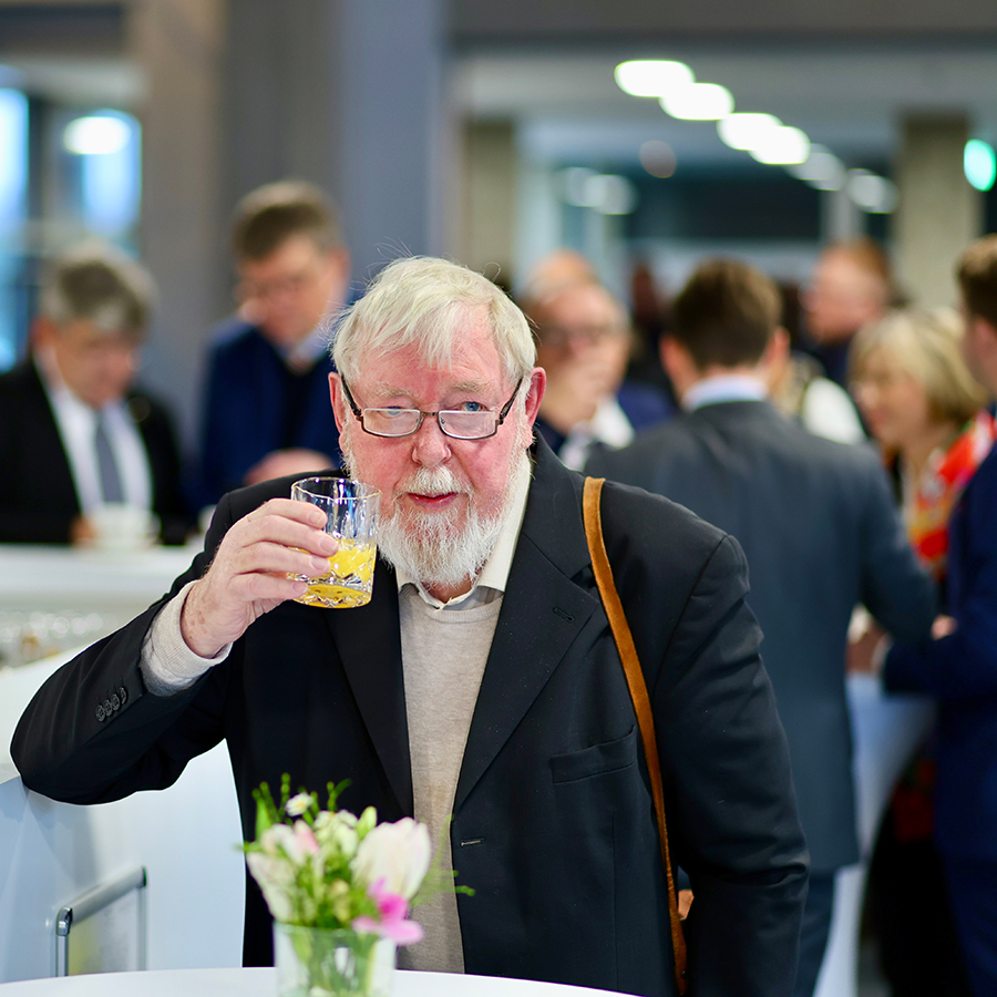 Michael Succow hält an einem Stehtisch mit einem kleinen Strauß Blumen ein Glas Orangensaft in die Höhe und blickt in Richtung Kamera