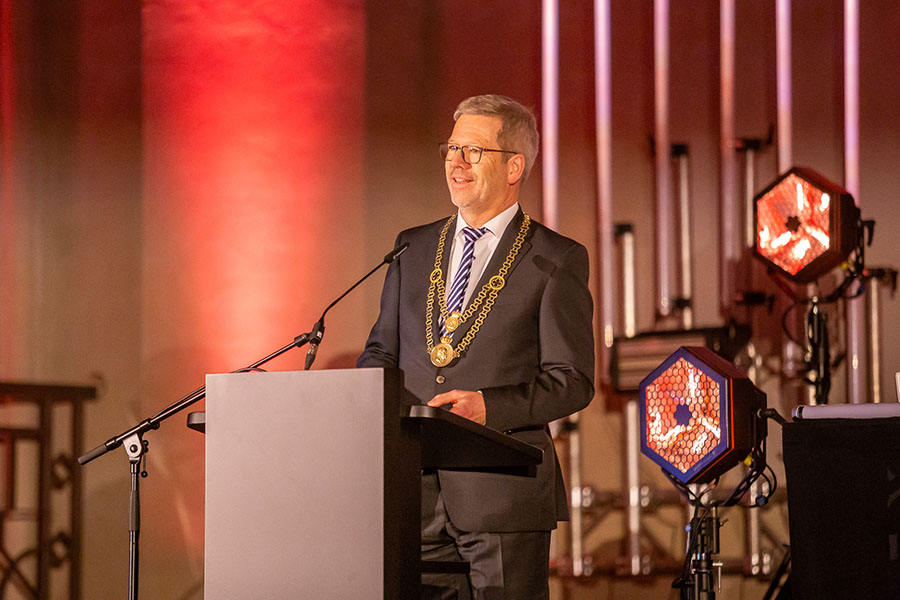 Greifswalds Oberbürgermeister Dr. Stefan Fassbinder bei der Begrüßung der Gäste im Greifswalder Dom. Er trägt seine Amtskette und steht an einem Pult mit Mikrophon. Im Hintergrund sind zwei Scheinwerfer zu sehen.