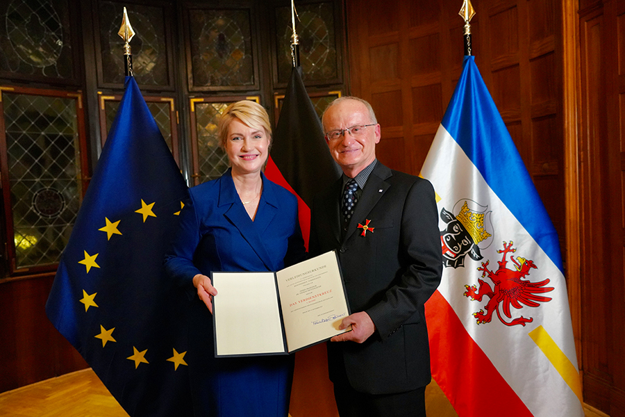 Prof. Dr. Thomas Mettenleiter und Ministerpräsidentin Manuela Schwesig halten die Verleihungsurkunde in die Kamera. Im Hintergrund sind die EU-, die Deutschland- und die Mecklenburg-Vorpommern-Flagge zu sehen.