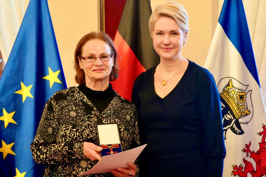 Die Ministerpräsidentin mit Rosita Mewis, die ihre Urkunde in der Hand und ihren Bundesverdienstorden hoch hält. Im Hintergrund sind die EU-, die Deutschland- und die Mecklenburg-Vorpommern-Flagge zu sehen.
