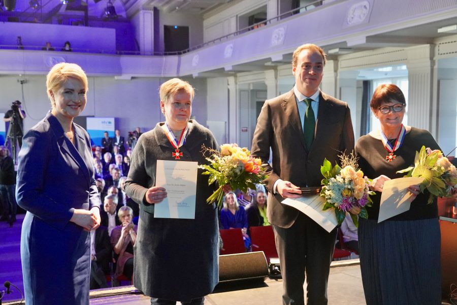 Die Ministerpräsidentin mit den Ausgezeichneten: Dr. Heike Müller, Jacob Uecker als Stellvertreter seines Vaters Günther und Sabine Kirton. Alle halten die Urkunden und Blumensträuße in ihren Händen. Dr. Heiko Müller und Sabine Kirton tragen ihre Verdienstorden um den Hals.