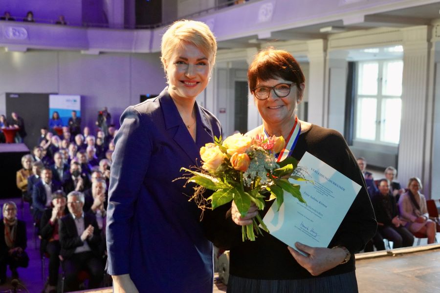 Ministerpräsidentin Manuela Schwesig mit Sabine Kirton, die einen Blumenstrauß und ihre Urkunde in den Händen hält.