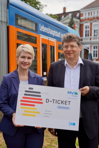 Ministerpräsidentin Schwesig und Minister Meyer halten gemeinsam eine vergrößerte Schautafel mit der Abbildung des Seniorentickets in MV hoch. Im Hintergrund ist eine Straßenbahn zu erkennen.